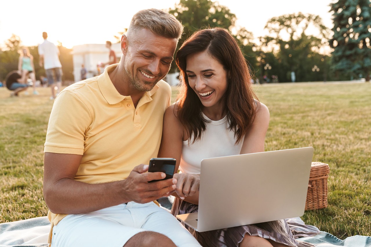 A couple on a picnic is smiling while sharing a smartphone screen.