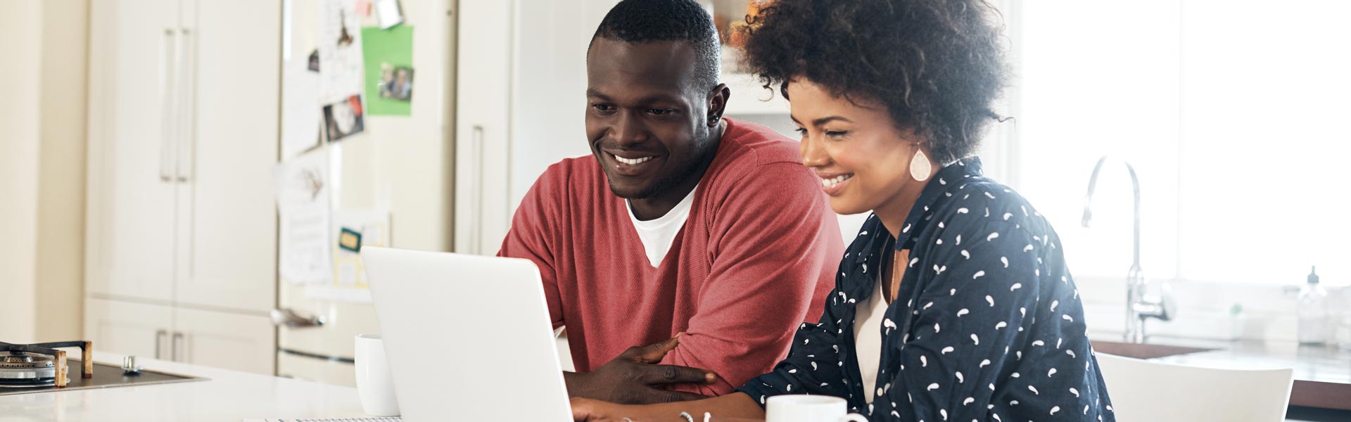 A couple sits at the kitchen table, looking at a laptop and smiling.