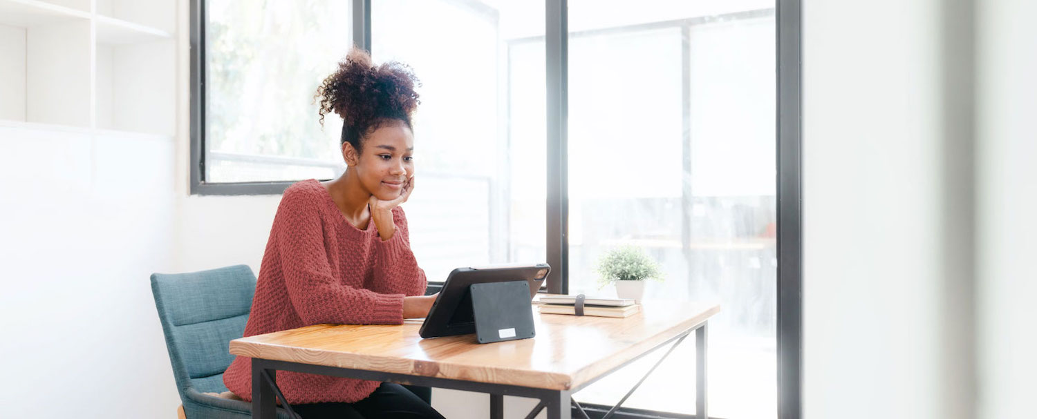 A woman sitting at a desk in an office reads from a tablet next to a small plant and pile of books. 