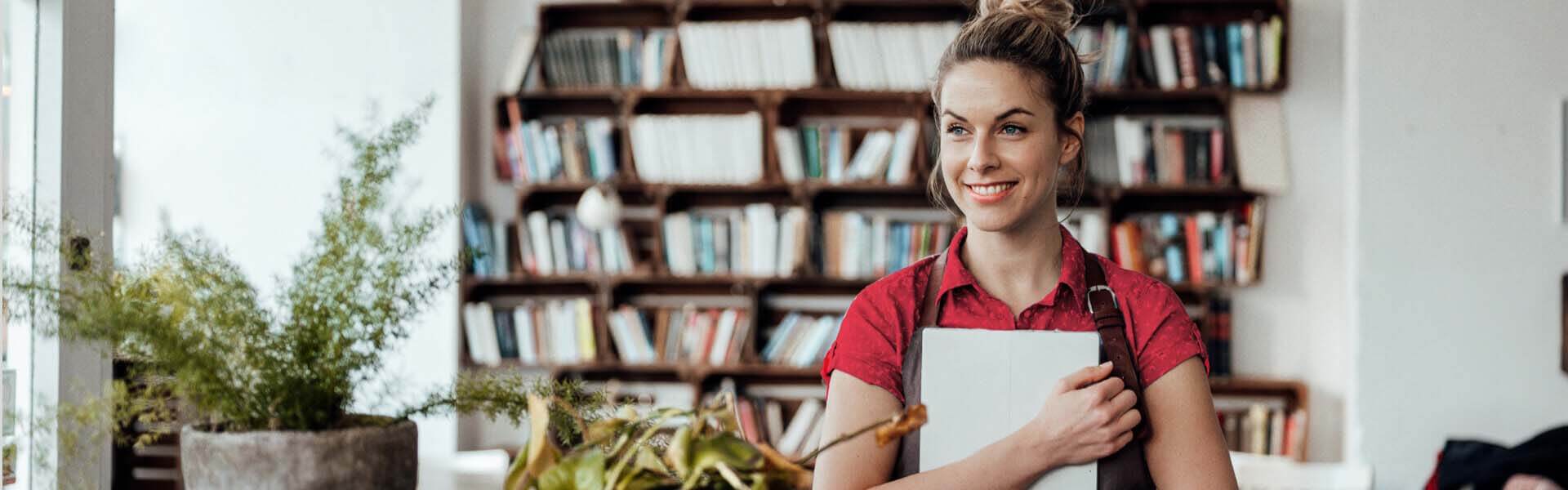 A young woman in a room lined with bookshelves hugs a notebook to her chest, smiling and looking confident.