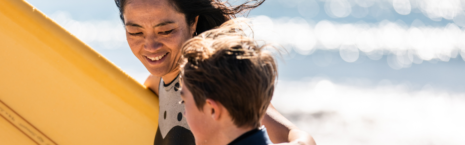 A mother is carrying a surfboard while walking next to her son.