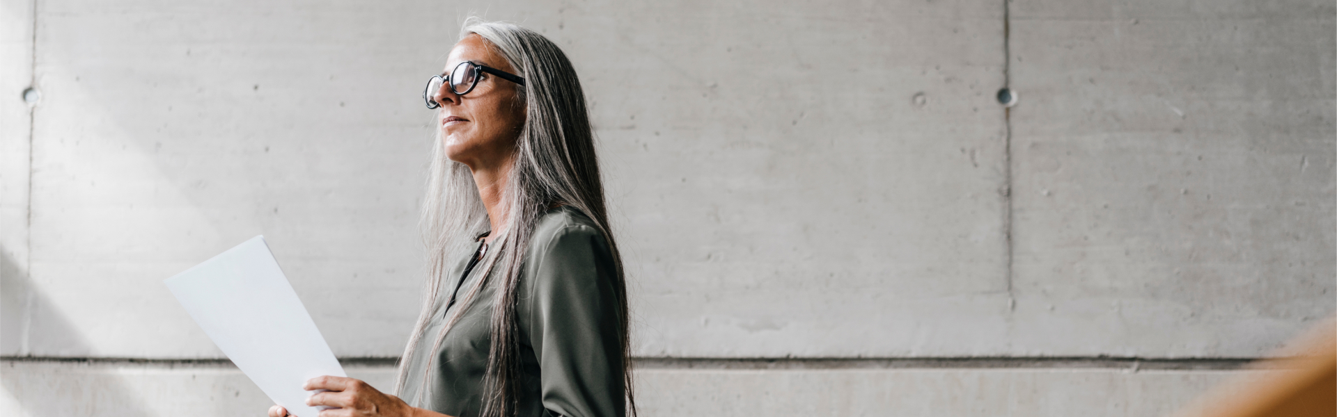 Lady in black glasses holds paperwork against concrete wall while pondering future.	