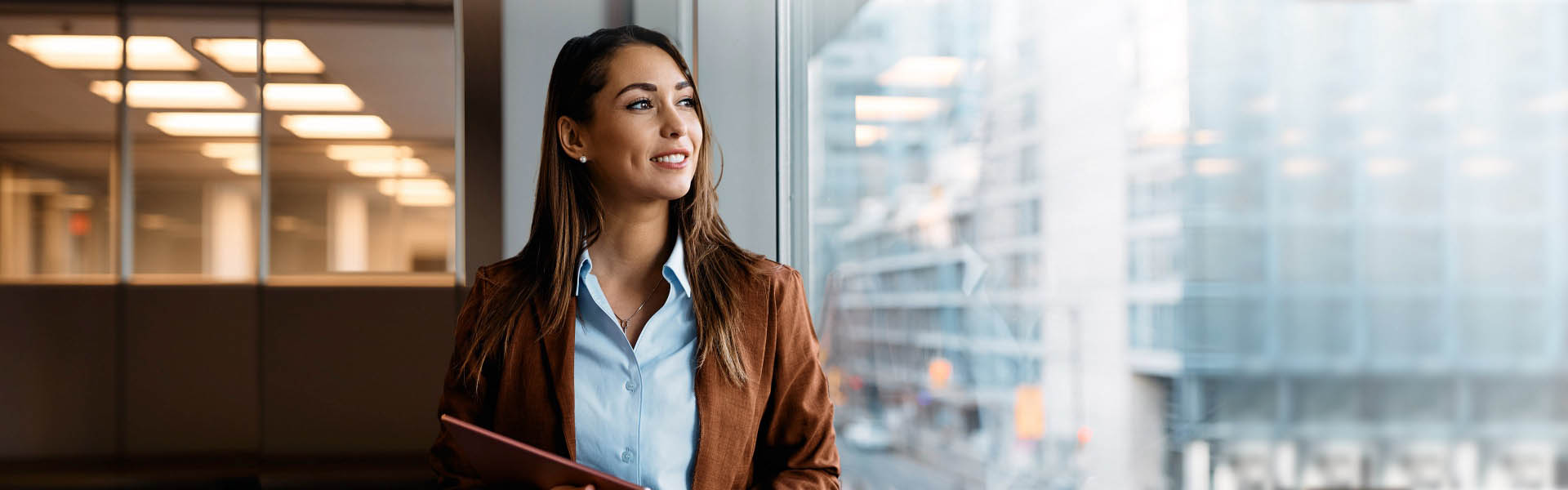 Smiling person looking out the window from an office inside a city building.  